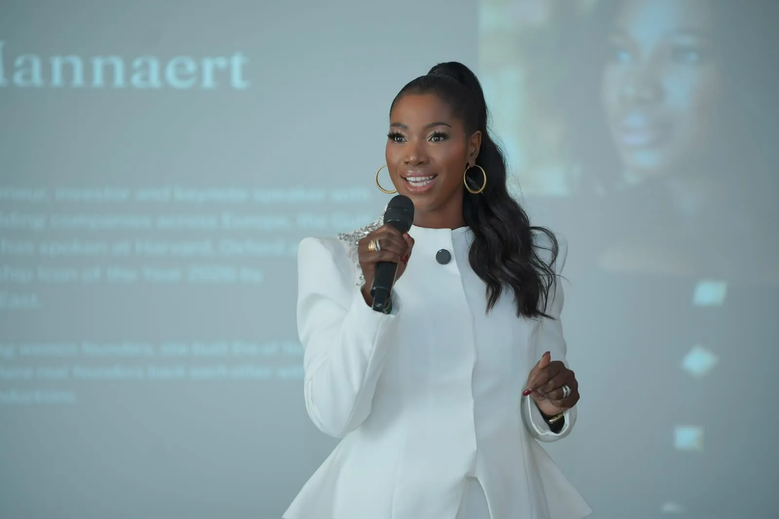 Ellen Mannaert speaking into a hand microphone, in a white jacket and gold hoop earrings, a projected slide with her name and portrait behind her.