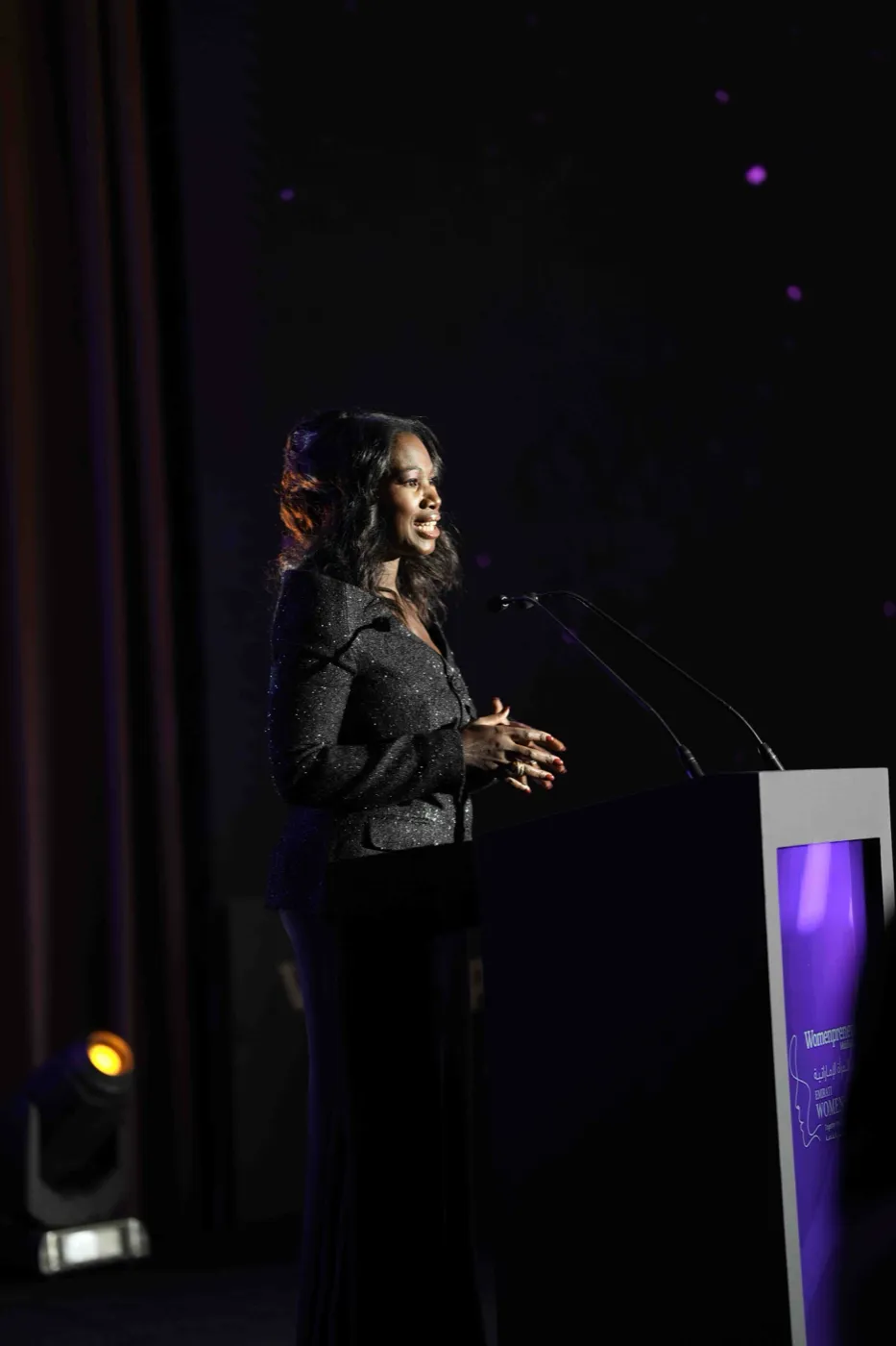 Ellen Mannaert speaking at a lit podium at the Womenpreneur Middle East Emirati Women’s Day Summit, a dark stage behind her.