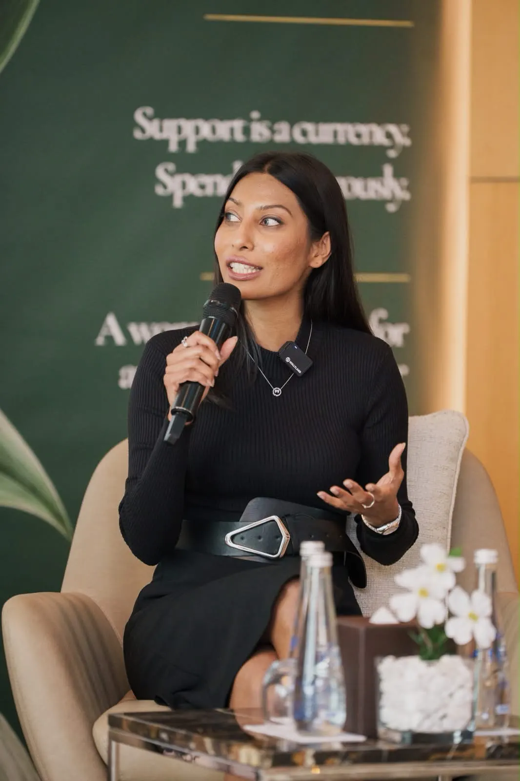 A panellist in black speaking into a microphone, a Wealth Room banner behind her.